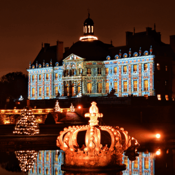 Illuminated Château de Vaux-le-Vicomte at night with a glowing crown sculpture and Christmas trees reflected in the water