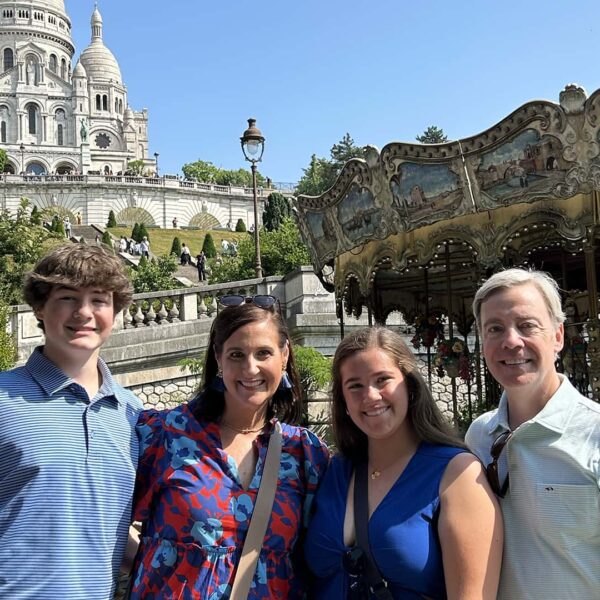 Family of four smiling by the Montmartre carousel with Sacré-Cœur basilica rising above them on a sunny private tour.