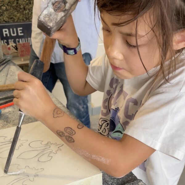 Young girl concentrating as she uses a hammer and chisel to carve a fleur-de-lis design into a stone block during a family workshop