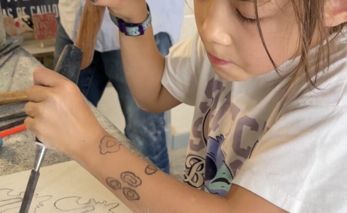 Young girl concentrating as she uses a hammer and chisel to carve a fleur-de-lis design into a stone block during a family workshop