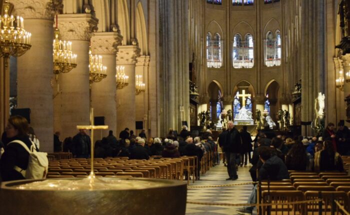Visitors sit and walk inside Notre-Dame Cathedral, facing the altar and golden cross beneath glowing chandeliers and stained-glass windows