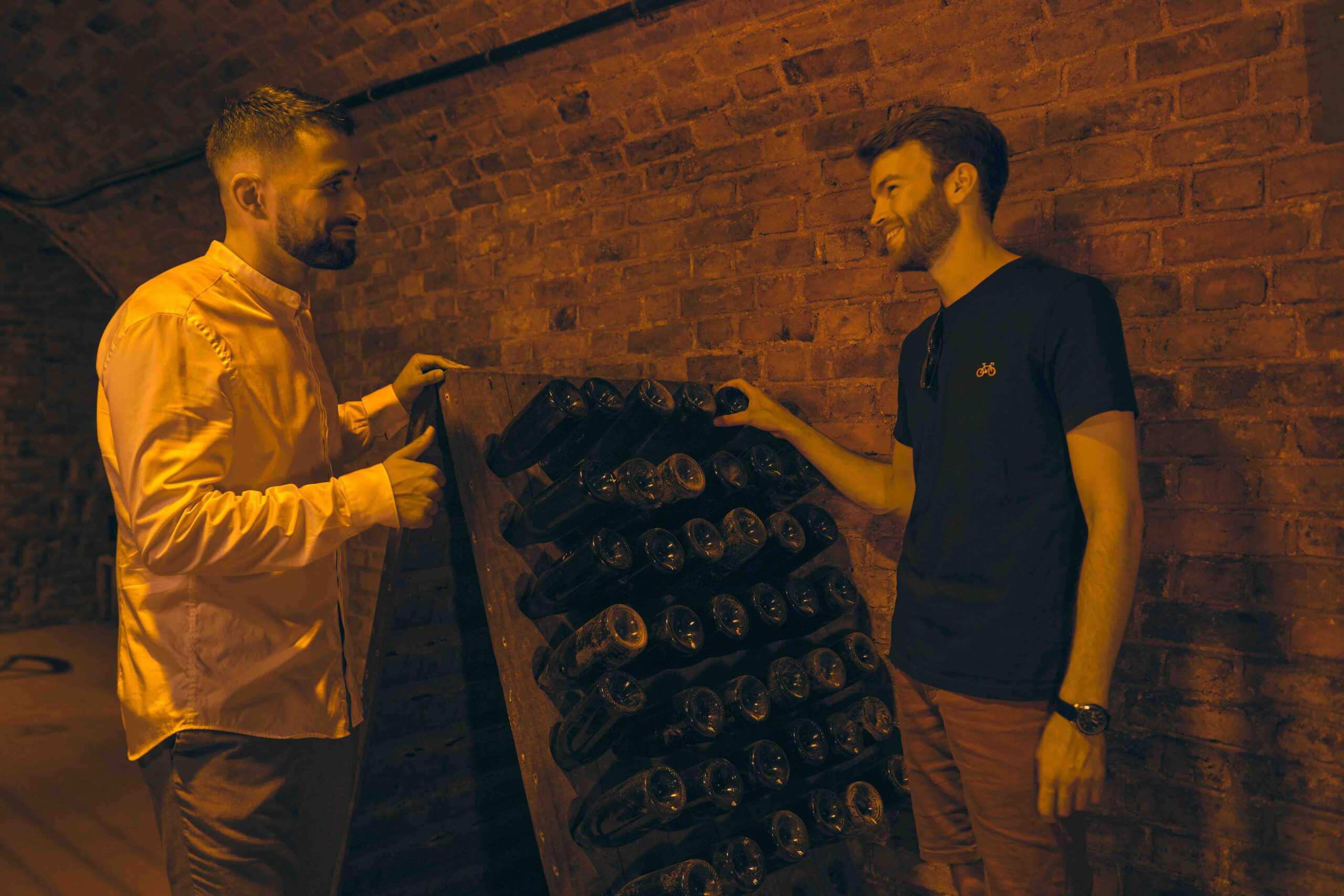 Learning riddling in Champagne cellars by train Two men chat beside a riddling rack in a Champagne cellar, holding dusty bottles while discussing the Champagne-making process.