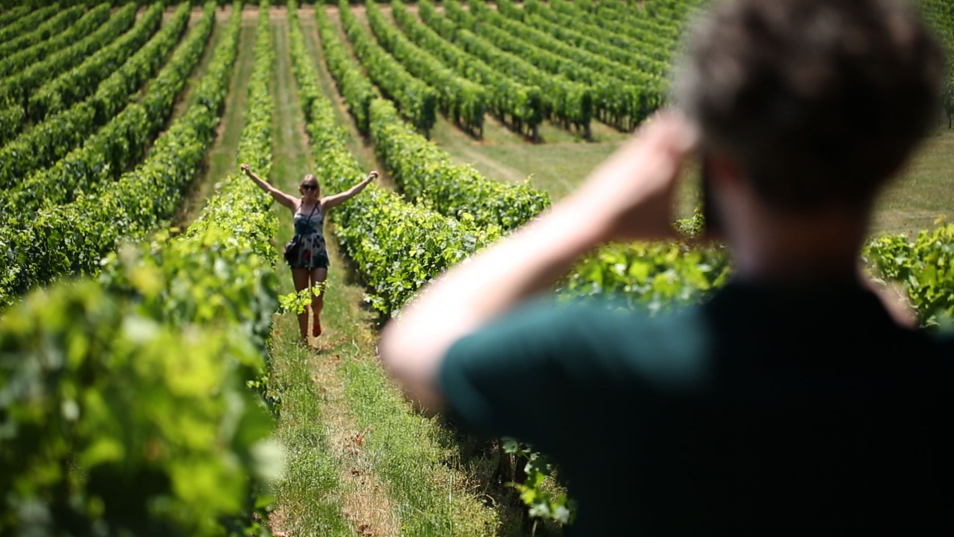 Woman joyfully posing between lush Champagne vineyard rows while a friend photographs her during a Champagne day trip from Paris