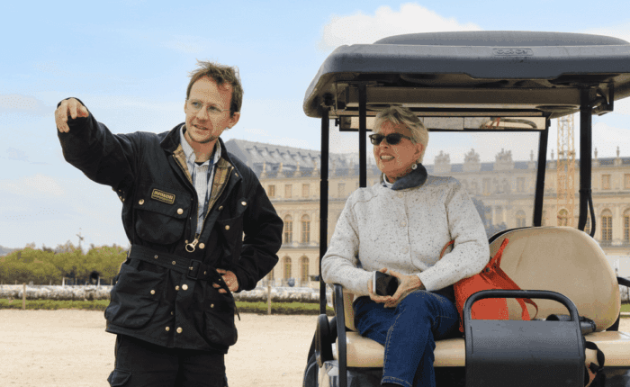 Guide standing beside a golf cart, pointing out sights to a seated guest with Versailles Palace in the background on a private tour.