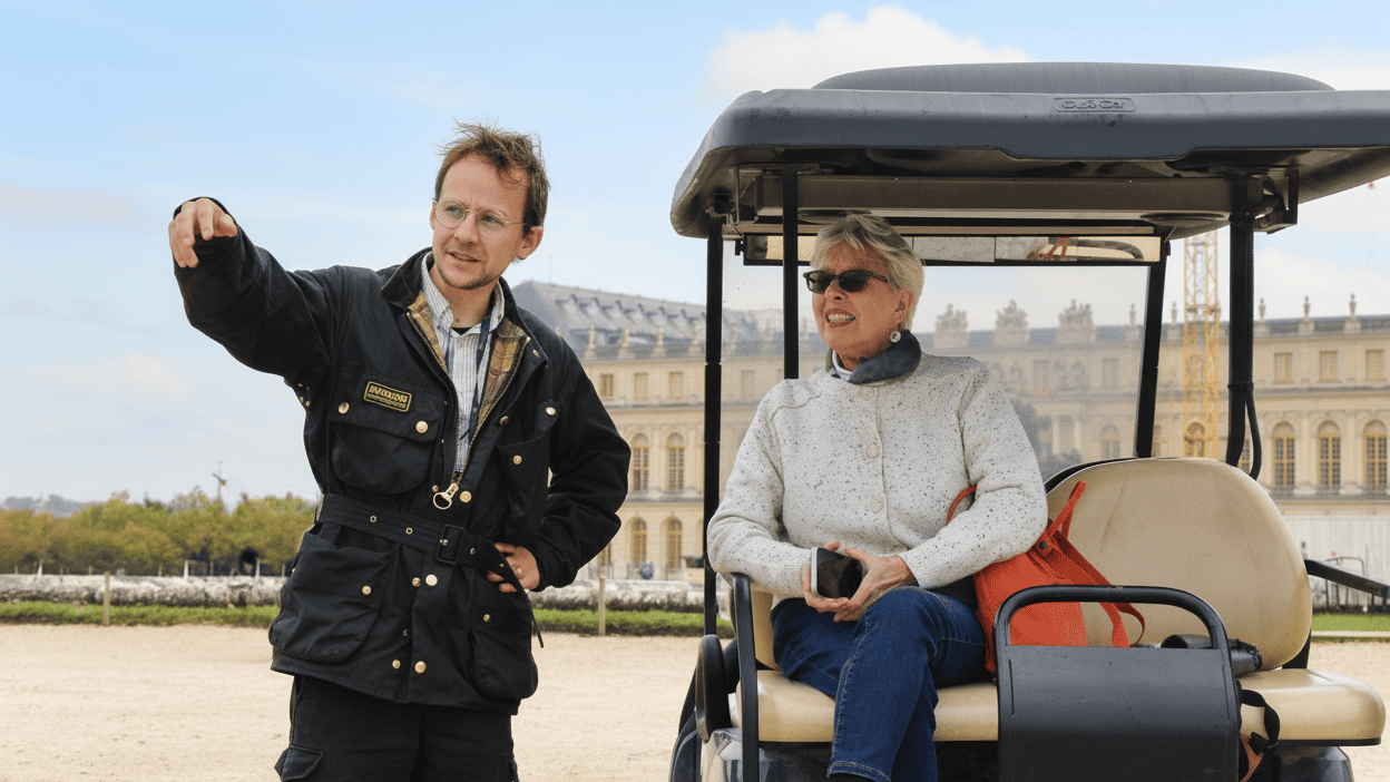 Guide standing beside a golf cart, pointing out sights to a seated guest with Versailles Palace in the background on a private tour.